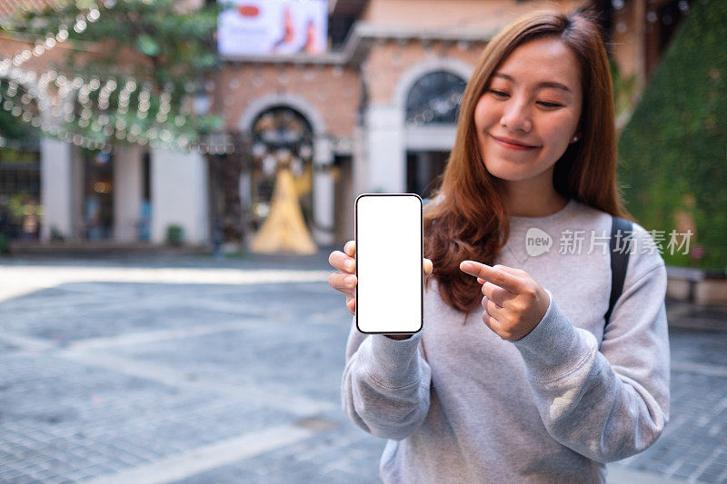 模拟图像的一个年轻的亚洲妇女持有和指着手指在户外的一个空白屏幕的手机