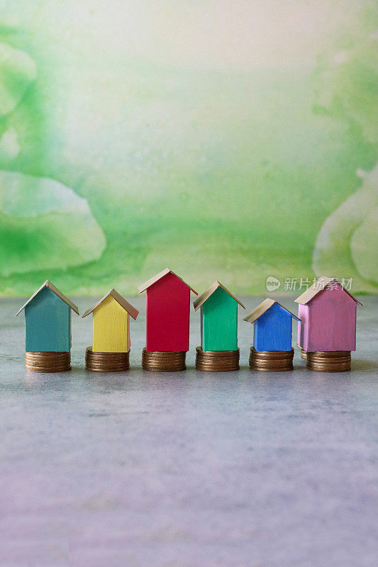 一排排彩色纸板房子的形象，五颜六色的海滩小屋矗立在成堆的硬币上，重点放在前景，斑驳的绿色背景，家庭财务，房地产和假日储蓄的概念