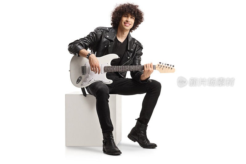 穿着皮夹克的年轻摇滚男歌手拿着一把电吉他坐在一个白色的立方体上