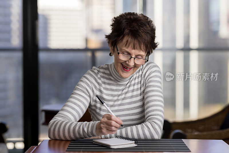 照片中，一位面带微笑的年长女性坐在桌旁，用笔在笔记本上写字