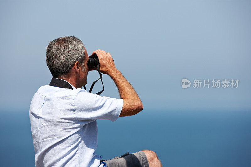 一个成熟的白人男人坐在悬崖顶上用望远镜看东西