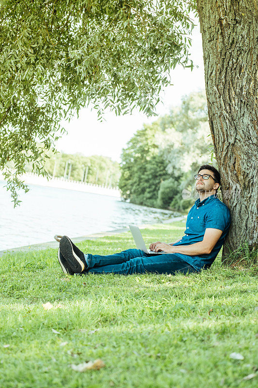 一个年轻人带着笔记本电脑坐在户外的草地上。远程工作的概念