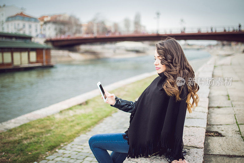 美丽的黑发女孩坐在城市河享受空闲时间使用手机和自拍。