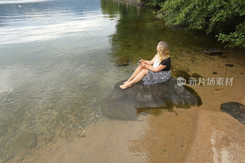 成熟的女人坐在淹没在水里的岩石上发短信