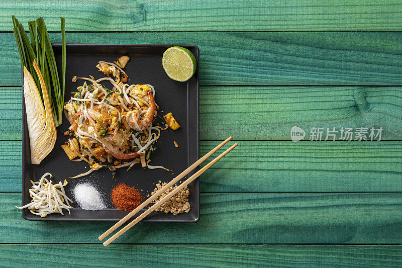 世界著名的鲜炒泰式虾仁泰式炒面放在一个方形的黑色陶瓷盘子上，筷子放在盘子的一角，桌子的背景是抽象的风化的绿松石色木面板。