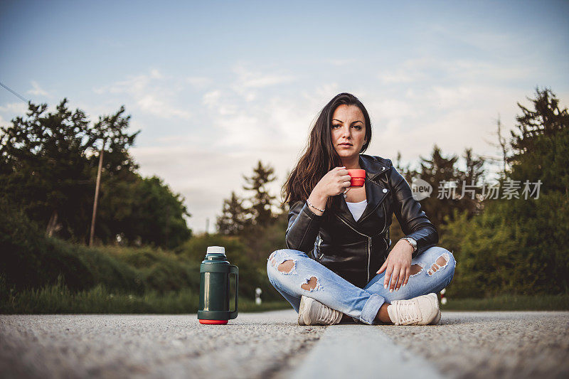 一个年轻的女孩正坐在柏油路上喝着咖啡-这个女孩正用一杯咖啡休息在路中间