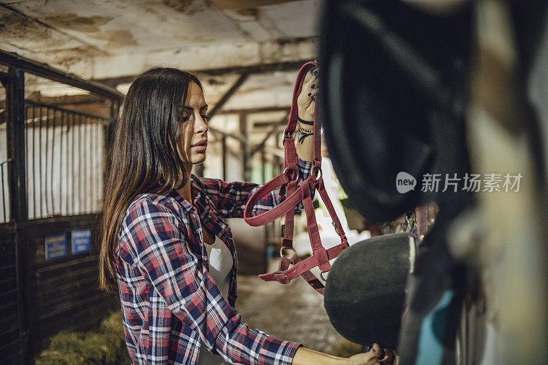 女人准备训练马，站在马厩里准备马鞍-一个年轻女人站在马厩里的马