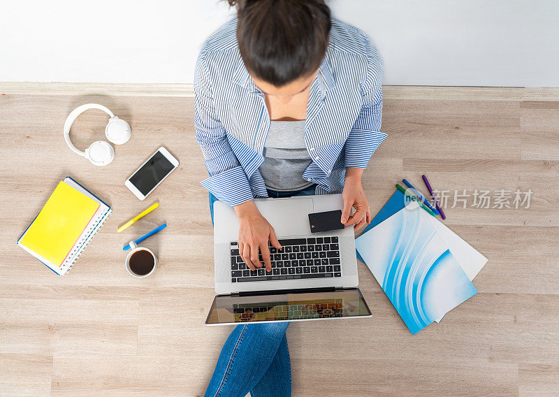 俯视图的照片，一个女人使用信用卡通过笔记本电脑在线支付，而坐在地板上。网上购物