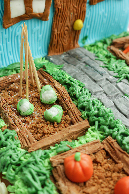 全帧图像的自制蛋糕设计为小屋和花园，黄油糖霜草和方糖糖霜通道，巧克力片种植的饼干屑土壤，方糖糖霜花椰菜和南瓜细节，聚焦前景