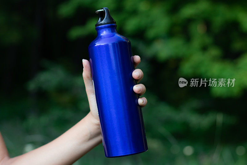 蓝色运动瓶的水在一个女性的手特写在自然或森林的模糊背景。在外面的街道上，手里拿着野营瓶，背景是绿色的。