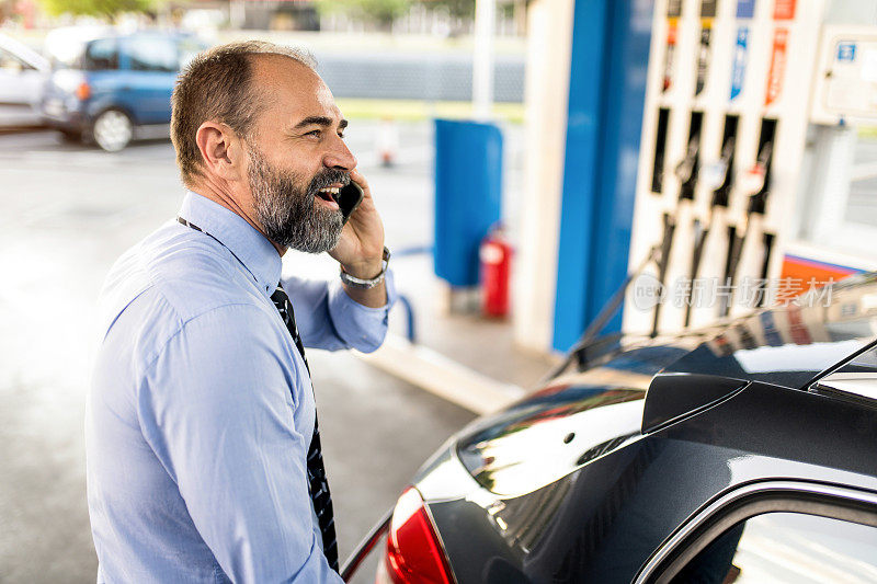 一个快乐的中年男人一边在加油站给车加油一边打电话。