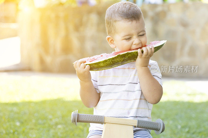 一个可爱的小男孩坐在花园里的自行车上吃着西瓜。夏天的乐趣和健康的食品，为年轻的孩子。