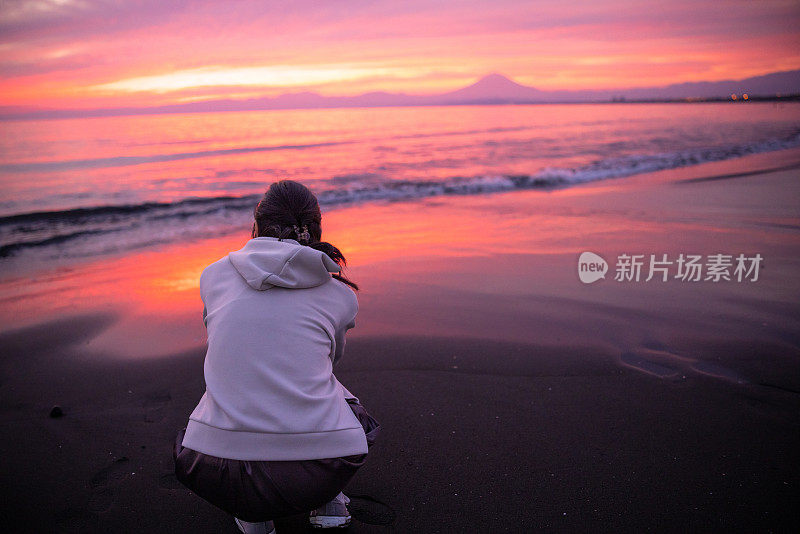 落日时分，坐在沙滩上看富士山的女子的后视图