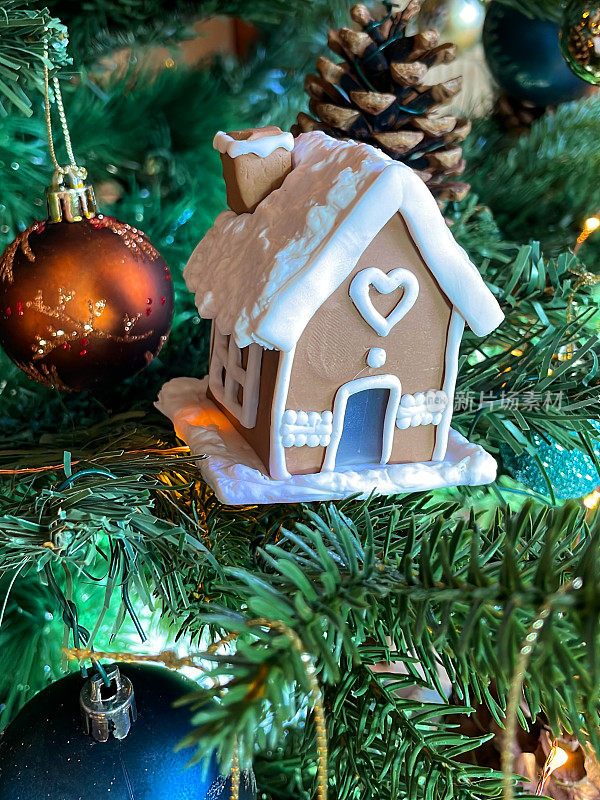 人造姜饼屋的形象，装饰有白色的糖霜装饰，松果和蓝色和青铜小玩意在人造圣诞树的树枝，云杉针叶，模糊的白色仙女灯背景