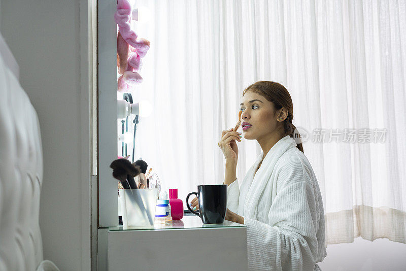 身穿白色浴衣的年轻拉丁女子正坐在梳妆台前化妆