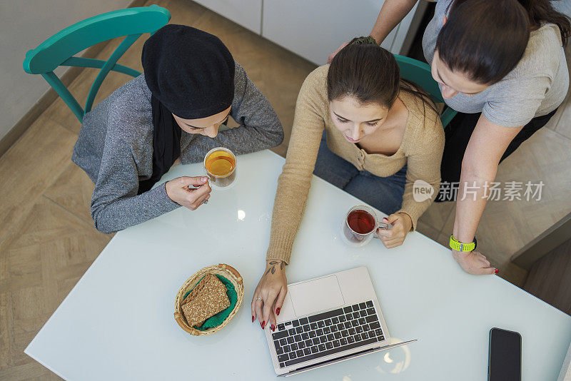 两位年轻女性，白人白人和中东穆斯林朋友，在厨房里用笔记本电脑一起工作。