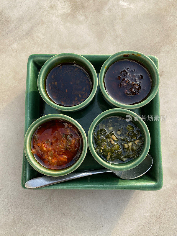 绿色方形的盘子上有四种蘸料，分别是辣椒油、酱油、西红柿和大蒜等辣味调料，从高空俯瞰