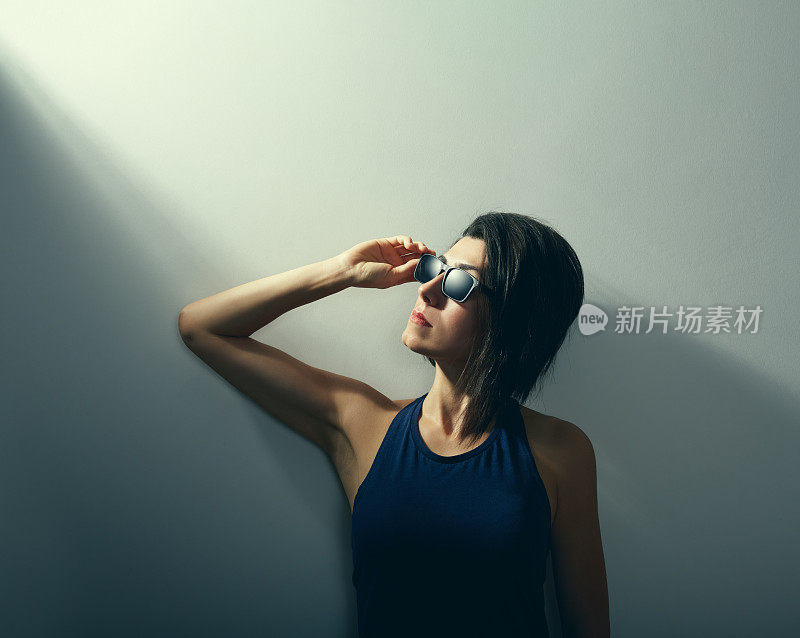 一个年轻的女人站在一个忧郁的环境的墙背景前，拿着她的太阳镜，看着光