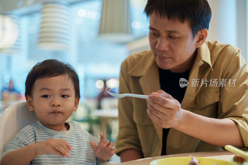 可爱的父亲一边喂他可爱的小儿子，一边和他一起在餐厅吃饭。