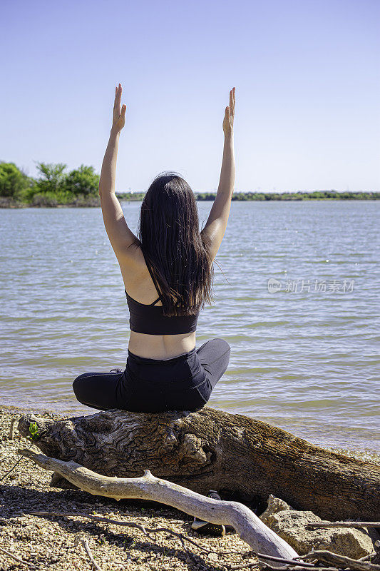 一名女子坐在一大块浮木上，手臂举在以湖为背景的岩石沙滩上