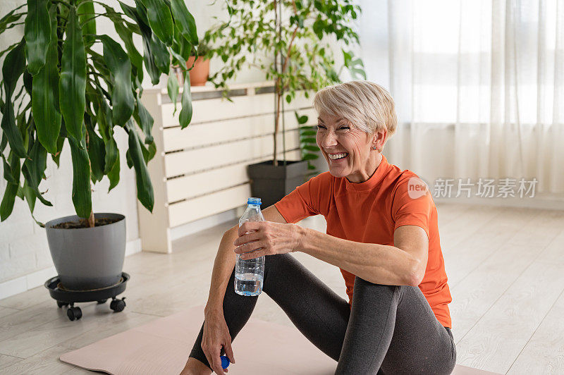漂亮的老女人运动后喝水。活跃的资深女性在室内练习瑜伽。老年人的运动