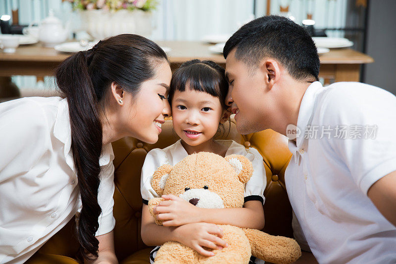 父母在房子里亲吻女儿，女儿抱着泰迪熊
