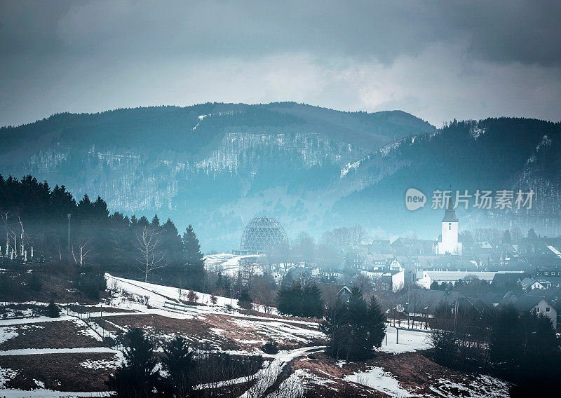 白色教堂在朦胧的雪山山谷与黑暗多云的天空。Winterberg,德国。