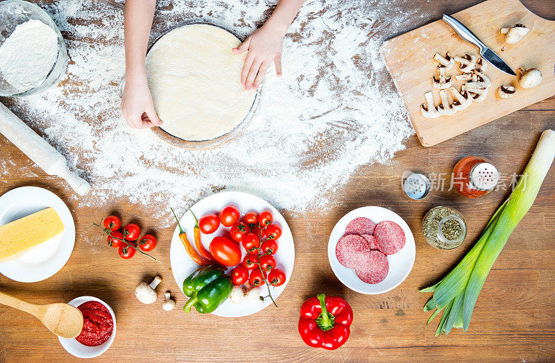 俯视图的孩子做比萨饼的配料，西红柿，意大利腊肠和蘑菇在木制的桌面上