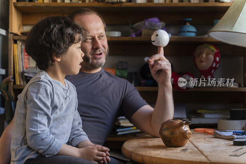 父亲是一个木偶师，在玩木偶，教他年幼的儿子存钱。