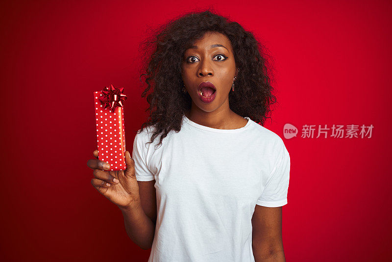 年轻的非洲裔美国妇女拿着情人节礼物站在孤立的红色背景吓了一跳，吃惊的脸，害怕和兴奋与恐惧的表情