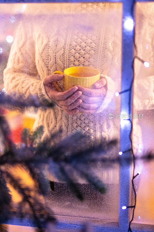 一个女人一边喝着热饮料，一边透过结霜的窗户看着窗外飘落的雪花