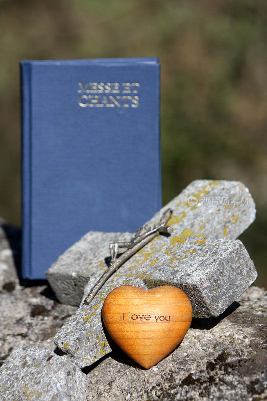 法国。2019年4月30日。橄榄木的心，上面写着“我爱你”，一本弥撒和歌咏书，一个十字架和一个在墓地墙上破碎的石头上的十字架。