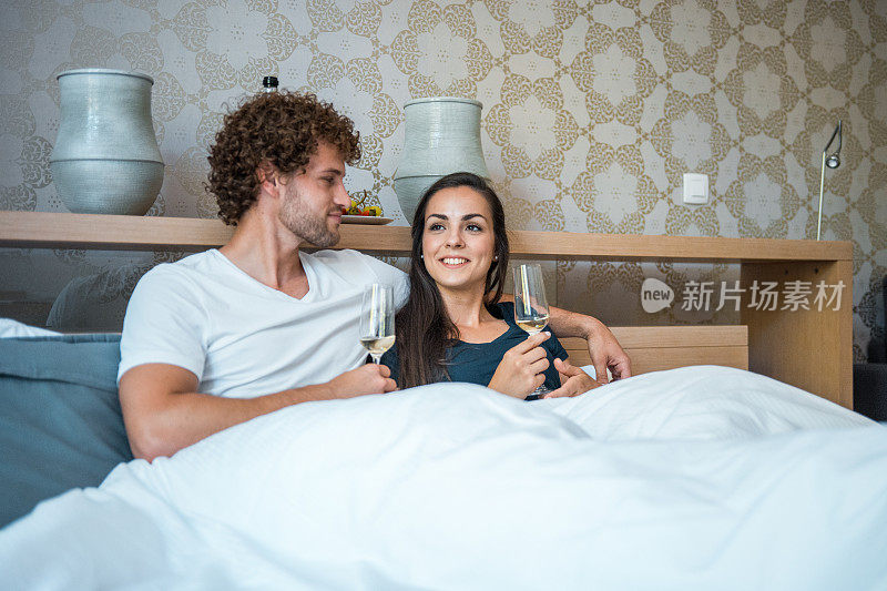 一对年轻夫妇躺在床上，喝着香槟庆祝搬家