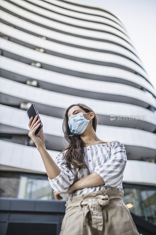 年轻女子站在城市街道上使用智能手机，脸上戴着防护面具。病毒大流行与污染概念。