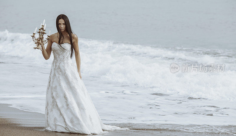 愤怒的年轻女子穿着婚纱，手拿烛台站在海边。