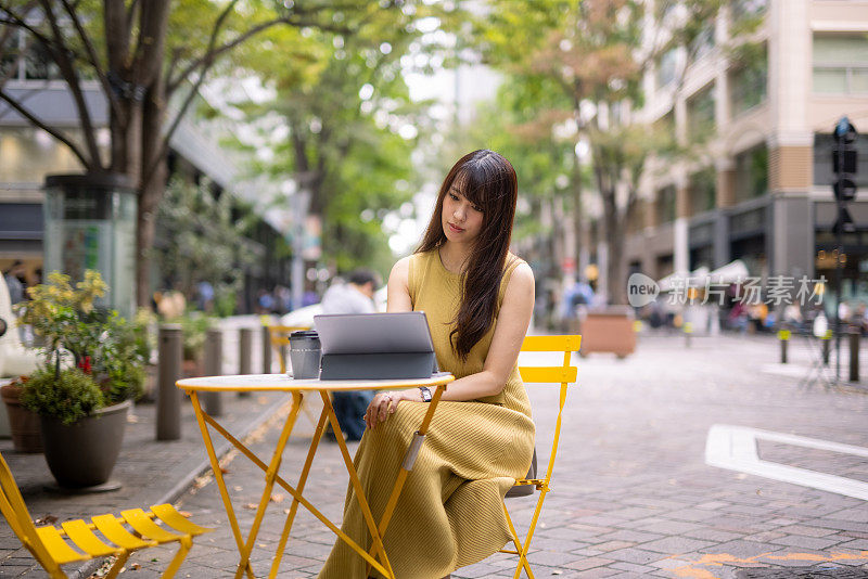 一名身穿黄色连衣裙的日本女子坐在城市街道的椅子上，正在使用电子平板电脑