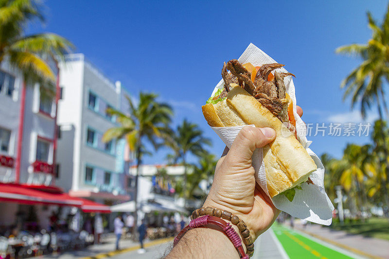 一个年轻的旅行男性享受他的假期，吃着古巴三明治在海洋大道前，南海滩，迈阿密，迈阿密，南佛罗里达，美国。