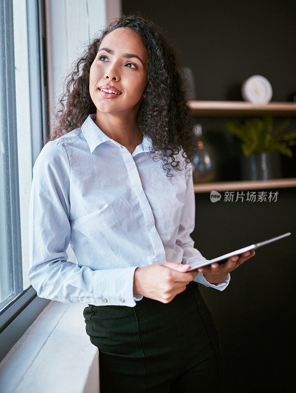 快乐的年轻的混血女商人拿着一个数字平板电脑，在办公室里浏览互联网。沉思和雄心勃勃的西班牙企业家使用技术作为一个自由职业者创业