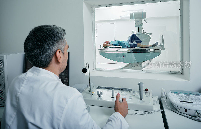 放射科医生坐在他的放射科医生办公室的保护窗后控制x光过程，而病人躺在放射成像室的x光机床上