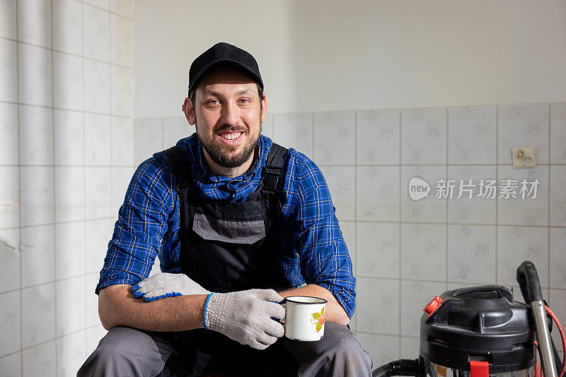 一个微笑的男人受雇在建筑工地工作的肖像。这个人正坐在一个正在拆迁的房间里休息，喝着咖啡和茶，这是翻新工作的间歇