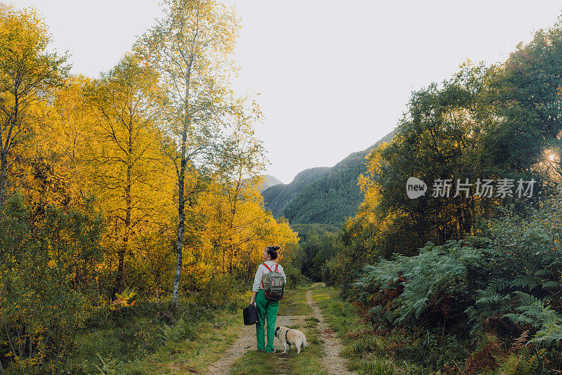 一位背着双肩包的女性带着她的狗在挪威山区的秋天森林里散步