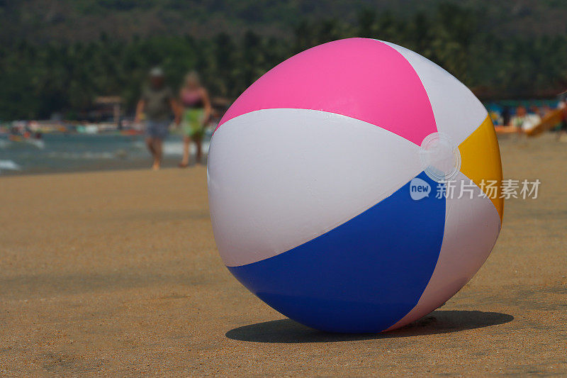水边沙滩上的沙滩球的特写图像，塑料，色彩鲜艳的儿童海滩玩具，退潮时无法辨认的游客走过破碎的海浪，林地背景，重点在前景