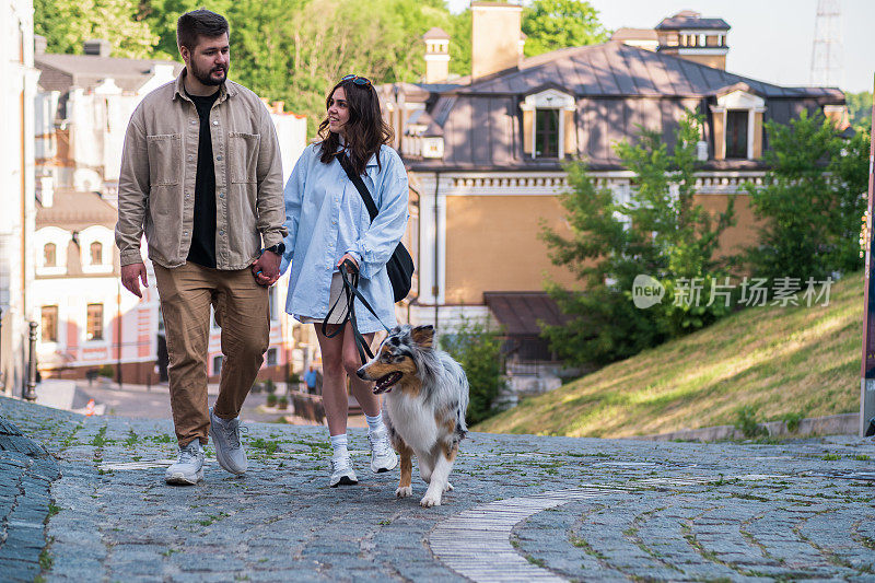 一对年轻夫妇和他们的澳洲牧羊犬在市区散步。幸福的男人和女人在城市里遛宠物，真正的生活方式