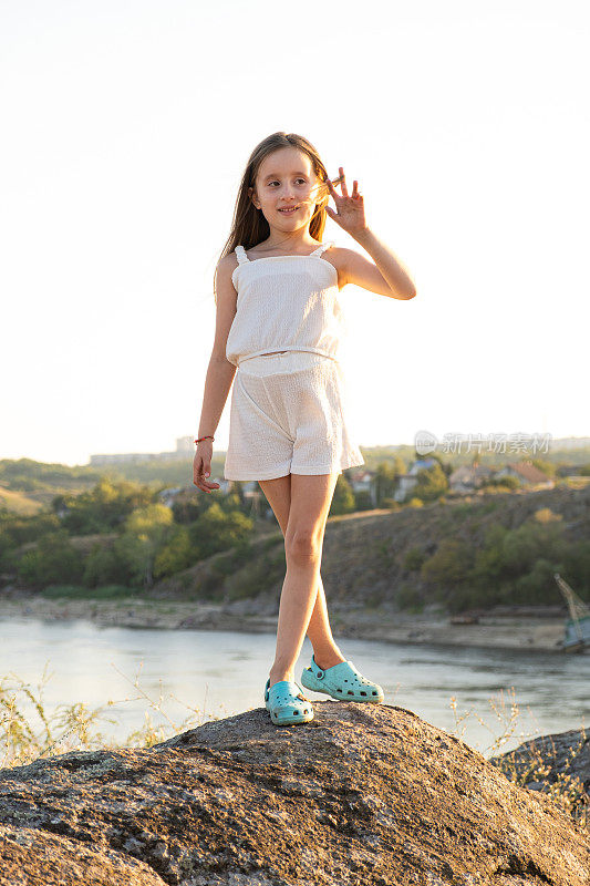 一个五岁的小女孩在大自然中站在一块石头上，背景是日落时的河流和天空。