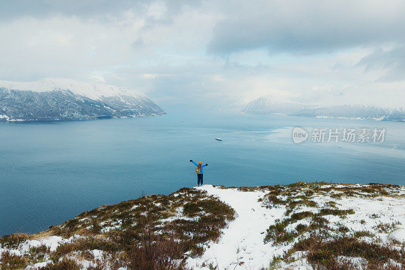 背着背包的女人站在山顶上举起双臂，从上面俯瞰冬日峡湾