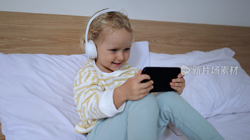 一个小孩半坐在父母卧室的床上。她把耳机戴在头上，里面放着轻松的音乐。女孩手里拿着手机玩游戏。
