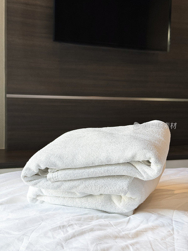 特写画面:白色羽绒被床罩上叠着白色酒店毛巾，床脚上叠着浴巾和手巾，壁挂式电视屏幕在浮动抽屉架上，重点放在前景