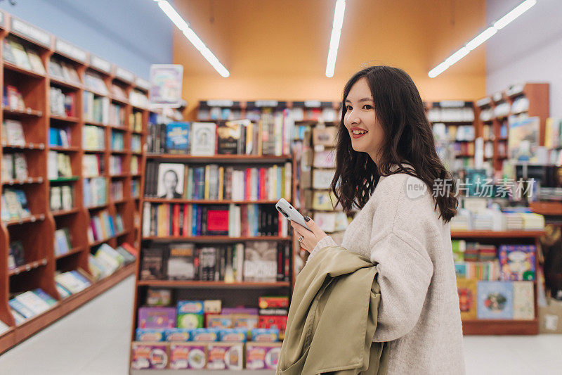严肃的青少年Z世代韩国女学生拿着手机，在大学图书馆使用智能手机上的app，浏览、聊天、上网咨询，心情愉快、开心、微笑。系列的一部分