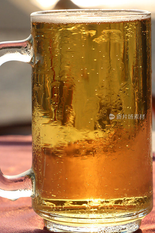全帧图像啤酒在玻璃啤酒杯覆盖在凝结与起泡的气泡形成泡沫的啤酒顶部的泡沫，马克杯品脱玻璃杯在沙滩酒吧户外桌子与桌布，重点在前景
