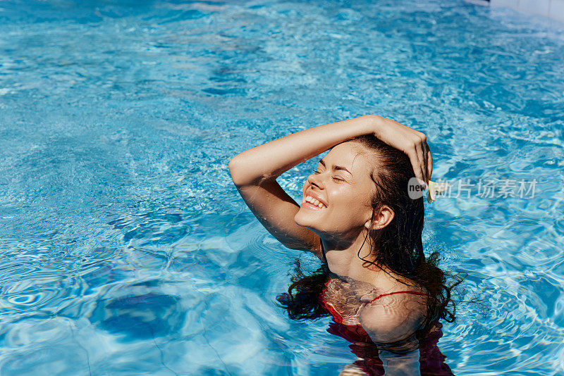 年轻女子在游泳池里穿着红色的泳装，很漂亮。在阳光下微笑，用手抚平头发，美丽而黝黑。